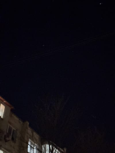 The image captures a nighttime outdoor scene in Ciorescu, Moldova. The upper two-thirds of the frame show a dark sky with scattered faint light points, consistent with stars, and multiple thin utility lines spanning horizontally across the central area. The lower left quarter depicts the upper section of a multi-story building, likely residential, with visible windows. At least two windows are illuminated from within, one of which contains vertical blinds. A deciduous tree, bare of leaves, occupies the lower-center and right portions of the frame, its branches silhouetted against the dark sky. No individuals or discernible actions are present.