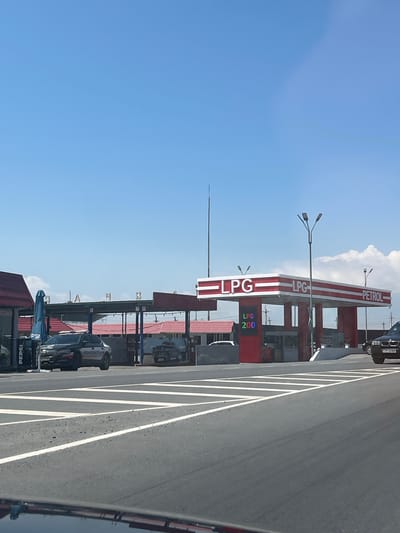 The image shows a roadside view of a petrol station. The station has a distinctive red and white striped canopy over multiple fuel pumps. Visible text on the canopy reads "PETROL" and "LPG" multiple times. A digital display shows "LPG 200". Several cars are parked or driving at the station. One car, a black sedan, is visible at the far right of the frame. Another car, a white sedan with writing on its side, is parked under a canopy. A black car is parked at a pump. A Pepsi branded umbrella is also visible. The scene appears to be in a bright, possibly sunny, daytime setting, with a clear blue sky and some scattered clouds. The road in the foreground is paved with white lane markings. The location is identified as Surenavan, Armenia.