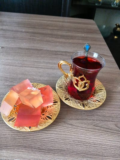 The image shows a close-up, eye-level view of a table setting with two golden decorative plates, one holding several pieces of cubed, translucent, pinkish-orange candy, and the other holding a traditional Turkish tea glass filled with dark red liquid. The tea glass has a decorative golden handle and is resting on a matching golden saucer. A small spoon with a blue embellishment is in the tea glass. The table appears to be made of wood with a visible grain. The background is slightly blurred, showing hints of dark furniture and possibly other decorative items, suggesting an indoor setting. The lighting appears to be natural, possibly from a window, casting soft shadows. The overall mood is one of a calm, perhaps leisurely moment, with the sweet candy and hot tea suggesting a break or a treat. There is no visible text in the image. The location context provided is Dzhiginka, Russia, which implies a domestic or hospitality setting within that region.