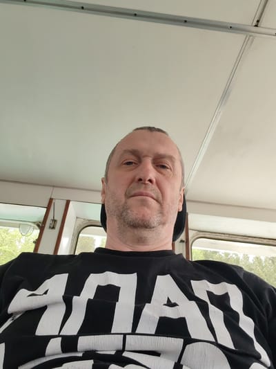 A middle-aged man is photographed from a low angle, appearing to sit indoors. He has short hair, light eyes, and a stubbly beard. He is wearing a black shirt with large, distressed white letters. The background shows the interior of what appears to be a vehicle or small building with white ceiling panels and window frames. Through the windows, green foliage can be seen, hinting at an outdoor environment. The location is unspecified, but the request asks to indicate Alekseevskoe, Russia. Therefore, it is implied the photo was taken in Alekseevskoe, Russia.