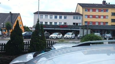 The image shows a street scene in Altdorf bei Nürnberg, Germany. In the foreground, the roof of a silver vehicle is visible. Behind it, a dark green fence runs horizontally across the frame. Beyond the fence are two tall, dark green evergreen trees. In the middle ground, several cars are parked in front of a building that houses a "Backhaus Fuchs" bakery and a "Metzgerei" (butcher shop), as well as "Obst & Gemüse" (fruit and vegetables). The building has a light-colored facade and several rows of windows. To the left of this building is an orange house with a dark roof and visible windows. To the right of the bakery building is a taller, yellow building with red shutters on its windows. The sky is overcast.
