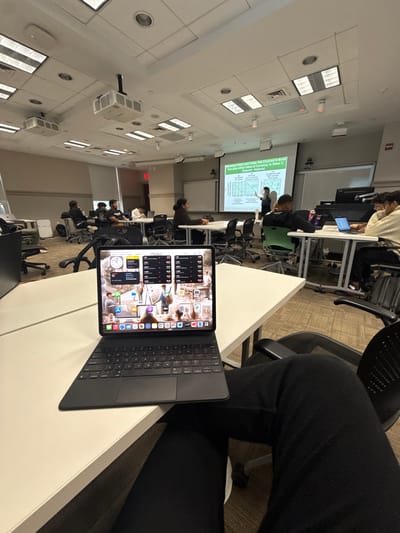 This image depicts a classroom in Wellesley, United States. In the immediate foreground, an iPad Pro with a black Magic Keyboard case rests on a white table, displaying its home screen with multiple widgets and app icons. The perspective is from a seated individual whose dark-colored pants and a partial white shoe are visible beneath the table.

In the mid-ground, several individuals, presumably students, are seated at white modular tables arranged throughout the room. Many students are observed looking at laptops or other devices on their desks. To the far left, an individual is visible seated at a desk with a computer monitor.

In the background, a person is standing at the front of the room, gesturing towards a large white projection screen. The screen displays a presentation slide with text prominently featuring "Monetary Policy and Trade, Not Covered in Book." Overhead, a ceiling-mounted projector casts the image onto the screen. The room features a white drop ceiling with rectangular fluorescent light panels, circular recessed lights, and visible speakers. An exit sign is illuminated on the left wall. The floor is carpeted. The scene suggests an educational setting, likely a college or university lecture or study session.