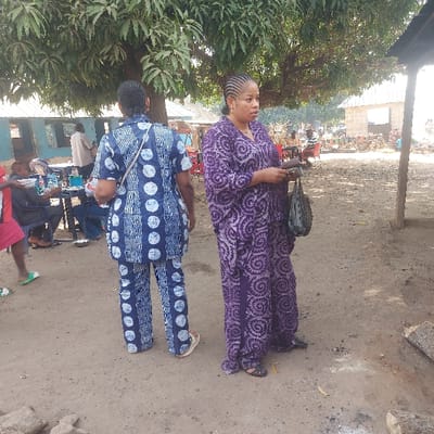 The scene depicts an outdoor gathering in Assai Sabongida, Nigeria, under bright daylight. In the foreground, two women stand on uneven, light brown dirt ground. The woman on the left is positioned with her back to the viewer, wearing a matching blue two-piece outfit (short-sleeved top and pants) adorned with a pattern of white circles and vertical wavy lines. A light-colored strap is visible across her right shoulder, and she wears light-colored sandals.
The woman on the right is shown in profile, facing towards the right side of the frame. She wears a long, loose-fitting purple garment with a distinct swirling pattern. Her hair is styled in dark braids. She holds a dark, elongated object in her right hand and a dark, possibly woven bag in her left hand. She wears dark sandals.
In the midground, under the shade of a large tree with green foliage, several individuals are seated at dark tables, appearing to eat or socialize. A person in a red shirt is partially visible on the far left. The background on the left features a long, low building with a blue exterior and multiple dark windows. To the right, a structure with a dark roof supported by a wooden post is visible. Further in the distance, a light-colored brick building and faint outlines of additional people or market displays can be observed. Scattered debris, including pieces of wood, are on the ground in the lower right foreground.