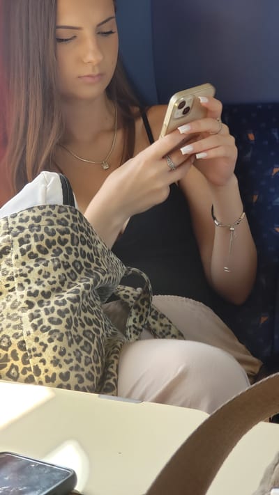 The image captures a young woman with long, brown hair, focused on her smartphone. She is seated, with a leopard-print bag on her lap and wearing a black top and light-colored pants. A delicate necklace with a square pendant adorns her neck, and she has several rings on her fingers and a bracelet on her wrist. The background suggests an indoor setting, possibly public transport, with dark blue seating and orange accents. The lighting is soft, casting gentle shadows and highlighting the textures of her clothing and accessories. The image does not provide any specific visual cues to confirm or deny the location being Považany, Slovakia.