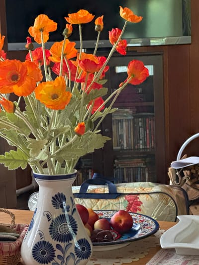 An indoor still life scene is depicted, likely in a living room or dining area, given the presence of a television, wooden cabinetry filled with media, and a wooden table. The foreground is dominated by a bouquet of vibrant orange poppies in a white ceramic vase with blue floral patterns. The vase sits on a lace-trimmed tablecloth. To the right of the vase, a decorative plate holds several fresh fruits, including peaches and possibly plums, alongside some dark chocolate pieces. A woven basket containing coasters is visible on the left side of the table. In the background, a dark wooden bookshelf or cabinet is filled with numerous books or DVDs. Above the shelf, a large flat-screen television is mounted. To the far right, a stack of firewood and a white ceramic pitcher are partially visible. The lighting suggests an indoor environment with natural light possibly supplementing artificial light. No people are present in the image. The scene appears to be set in the city of Газета Труд Снт, Russia.