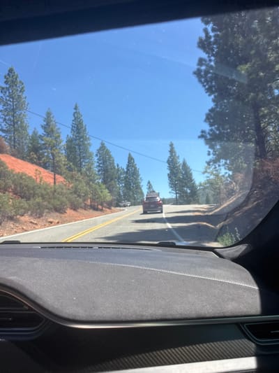 The image shows a view from inside a vehicle driving on a rural, two-lane road on a clear, sunny day. The road is paved with yellow double lines marking the center. The surrounding environment consists of a reddish-brown embankment with sparse vegetation on the left, and a dense forest of pine trees on the right. Several other vehicles are visible on the road ahead. The dashboard of the vehicle from which the photo is taken is partially visible in the foreground. There is no identifiable information to suggest the location is in White Hall, United States.