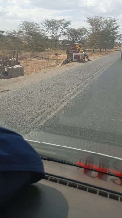 A roadside fruit stand with a man sitting nearby. The stand displays colorful fruits and vegetables. Behind the stand, the landscape features sparse, bare trees, suggesting a dry climate. A paved road runs in front of the stand, seen from the perspective of a vehicle's dashboard. The dashboard includes a blue object and partially visible red writing. The scene suggests a rural or developing area.