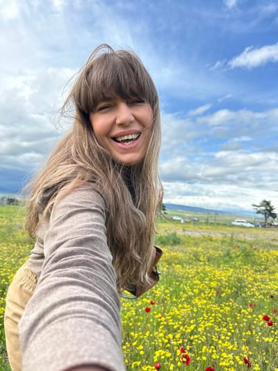 A smiling woman with long brown hair takes a selfie in a field of yellow and red flowers. The background consists of a blue sky with white clouds, a distant landscape with green fields, trees, and a few buildings. The photo appears to be taken at an angle, with the woman's head tilted. The exact location in Marneuli, Georgia, cannot be determined without more specific visual information.