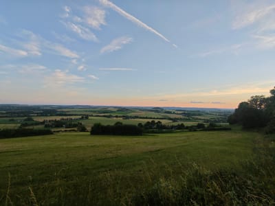 The image captures a panoramic view of a rolling landscape under a twilight sky, characteristic of the Teutoburg Forest region near Schieder-Schwalenberg, Germany. The foreground is dominated by a grassy field, with tall, dry grasses and wildflowers in the lower right corner suggesting a natural, untamed environment. Beyond this, the land descends into a valley dotted with trees and small clusters of buildings, indicating a rural settlement. The middle ground features a patchwork of cultivated fields in various shades of green and yellow, interspersed with dense green trees and hedgerows. In the distance, a hazy horizon reveals more undulating hills and hints of human presence, including wind turbines, a common sight in this part of Germany, and the soft glow of sunset colors in the sky. The sky itself is a gradient of pale blue, transitioning to warm oranges and pinks near the horizon, with thin, wispy clouds and a prominent contrail streaking across the upper portion. The overall scene evokes a sense of tranquility and vastness, typical of the German countryside at dusk. There are no people or animals visible in the image.