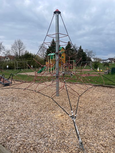 This image displays a vacant outdoor playground situated in Holtzheim, France, under an overcast sky. The foreground is covered with light brown wood chips. A prominent climbing net structure occupies the central and right portions of the frame, consisting of a vertical silver-grey metallic pole capped with a red cone. From this central pole, black and reddish-brown ropes extend outwards, forming a web-like arrangement, anchored to the ground by various metallic fasteners and a tensioning mechanism visible in the lower right.

In the midground, behind the climbing net, additional playground equipment is visible. This includes a multi-level play structure, predominantly yellow/tan and green, with a green slide. To the left of this structure, a swing set with two swings is present. To the right, two white swan-shaped spring riders, each with a red beak, are positioned on a red safety surface. Sections of green grass are interspersed between the various play areas. The background features a mix of bare deciduous trees and evergreen conifers, behind which several residential buildings with light-colored facades and dark roofs are observable. No individuals are present in the scene.