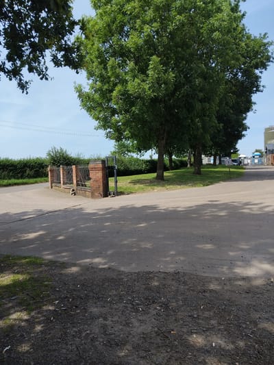 This media file shows an outdoor scene in a semi-rural setting, likely Stafford, United Kingdom. The viewpoint is at a low angle, looking up towards trees and the sky. A paved pathway runs diagonally through the frame, bordered by grass on the left and a rough, gravelly verge on the right.
The left side of the image is dominated by lush green trees, with dense foliage obscuring much of the sky. In the middle ground, a brick structure with a metal gate and fencing is visible. Behind this, a neatly trimmed hedge forms a boundary. Further back, a row of trees lines a grassy area, and beyond that, some industrial or commercial buildings with white roofs and tanks can be seen under a clear blue sky. A circular sign with the number "5" is affixed to a post amongst these trees.
The right side of the image features the paved path and the rough verge, which is littered with cigarette butts and small stones. Shadows from trees are cast across the path, indicating a sunny day, likely late morning or early afternoon. There are no people visible in the frame, but the presence of the pathway and buildings suggests this might be an area with some human activity or access. The overall impression is one of a well-maintained, yet somewhat untidy, outdoor space on a bright day. The text "5" is visible on a sign.