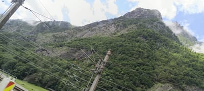 This image shows a view of a mountainous region in Golling an der Salzach, Austria. The foreground is dominated by numerous overhead power lines and a support pole, suggesting proximity to a railway line or an area with significant electrical infrastructure. The power lines stretch diagonally across the frame, creating a strong linear element.
Behind the infrastructure, a steep, densely forested mountainside rises dramatically. The trees are a vibrant green, indicating a lush summer or late spring landscape. Rocky outcrops and scree slopes are visible through the foliage, revealing the rugged terrain. In the upper portions of the mountain, patches of mist or low-lying clouds cling to the peaks, adding a dramatic and atmospheric quality to the scene. The sky above is partly cloudy, with bright white clouds against a blue backdrop. The overall impression is one of natural beauty and a wild, untamed landscape juxtaposed with man-made electrical systems. There are no people visible in the image.