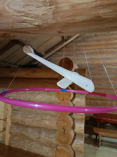 The image shows a wooden mobile hanging from a wooden beam ceiling, likely in a rustic cabin or home. The mobile consists of a white wooden airplane with wings and a tail, suspended by thin wires. Below the airplane, a large, bright pink hula hoop is also suspended, with blue tape wrapped around a section of its circumference. The background consists of wooden log walls, indicating an interior setting. The overall aesthetic is simple and handcrafted. The specific location in Марфино, Russia, is not identifiable from the visual information present in the image.