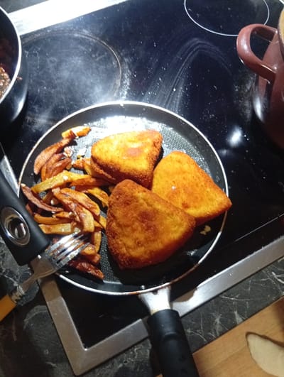 The image displays a close-up, high-angle view of a kitchen setting in Domsühl, Germany. The central element is a black non-stick frying pan, positioned on a black ceramic hob. Inside the pan are three golden-brown, triangular, breaded items, consistent with fried cheese or a similar cutlet, alongside a pile of golden-brown, irregularly cut fried potato pieces. A silver-colored fork with a yellow and black handle is resting within the potato pile. To the upper left, a second black pan is partially visible, containing dark, stew-like food remnants, with visible steam. To the upper right, a brown, handled ceramic pot is partially visible. The black ceramic hob shows some smudging and light reflections, and steam is evident above the cooking surfaces. The countertop underneath the hob is dark with light, marble-like veining. A silver-colored metallic edge is visible along the lower right side of the counter, with a light-brown wooden surface partially visible beyond it. No individuals are present in the scene.