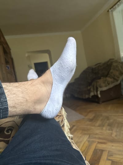 The image is a close-up shot of a person's right foot and leg, wearing a white ankle sock and dark jeans. The foot is positioned as if resting on another person's lap, which is covered in dark fabric. The background shows a room with wooden floors and a sofa covered with a patterned throw. The lighting suggests an indoor setting, possibly a living room. There is no visible indication of the location being in Tumanyan, Armenia.