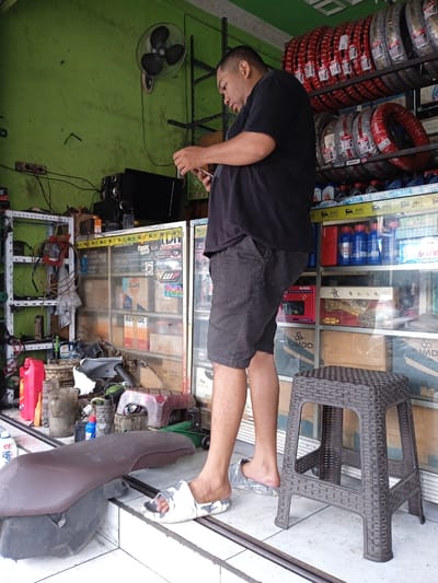The image depicts a man standing in what appears to be an automotive repair shop in Kauman, Indonesia. The shop's interior features a cluttered environment with various automotive parts and supplies.
The man is wearing a black t-shirt, gray shorts, and sandals. He is holding a mobile phone and appears to be focused on it.
Behind him, shelves are stacked with tires, motor oil, and other automotive products. A display case filled with automotive parts and accessories is also visible. To the left, there is a cluttered workstation with various tools and equipment. The walls are painted green, and a ceiling fan is mounted in the upper left corner.