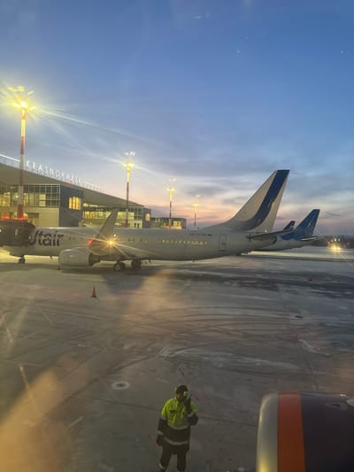 The image depicts an airport apron scene at Krasnoyarsk International Airport (Емельяново), Russia, during dusk or dawn. The sky transitions from deep blue at the top to hues of orange and pink near the horizon. The concrete apron below is marked with tire tracks and appears to be covered in a thin layer of ice or compacted snow.
In the mid-ground, a white Utair Boeing 737 aircraft, tail number RA-73145, is prominently parked. Its right engine cowling shows a bright light emanating from within or behind it. Further back, two additional white aircraft, sharing a similar blue-on-white tail fin design consistent with Utair, are partially visible.
To the left, a modern glass-fronted airport terminal building, displaying the illuminated text "KRASNOYARSK," is visible. Multiple tall light poles with bright yellow-white lights illuminate the terminal and apron area. An orange traffic cone is positioned on the apron near the front of the Utair aircraft.
In the lower central portion of the image, an airport ground crew member stands on the apron, wearing a high-visibility yellow and black jacket with reflective stripes, dark pants, and ear protection. The individual is holding a communication radio to their mouth, appearing to be in conversation. The photograph is taken from an aircraft window, with a portion of the observer's aircraft engine or wing visible in the bottom right corner.