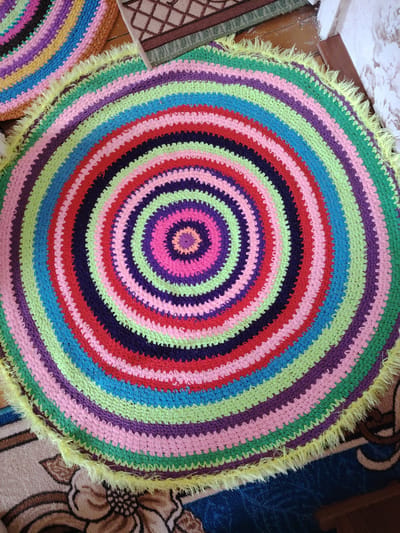 This image presents a vibrant, circular, handmade crocheted rug, prominently displayed on a floor in what appears to be an indoor setting. The rug features a striking pattern of concentric rings, crafted in a rich palette of bright colors including various shades of green, blue, pink, purple, and red. A distinctive detail is the fluffy, bright yellow fringe that outlines the entire circumference of the rug.
In the top-left corner, a fragment of a second, similar multicolored crocheted rug is visible, suggesting a coordinated set or multiple handmade pieces. The floor beneath and around the main rug is covered by other patterned carpets or rugs; one section visible in the background is a brown and beige carpet with a traditional floral or geometric design, while another area shows a blue carpet with darker patterns. Parts of a wooden floor or surface are also discernable.
No individuals are present in the image, and no specific activity is occurring. The photograph captures a static arrangement of home decor, illuminated by what appears to be bright, ambient daylight. The overall impression is one of handcrafted warmth and cheerful aesthetics. There is no visible text within the image. The location context for this scene is Yanaul, Russia.