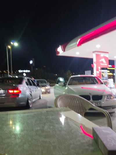 The image depicts a nighttime scene at a gas station in Güzelbahçe, Turkey. Several cars are visible, queuing up for service. In the foreground, a light-colored car is partially visible, with its red taillights illuminated. To the left, a white car with a red and white gas station sign overhead is prominent. The gas station's canopy features bright pink neon lighting. The overall impression is of a typical, busy night at a gas station, with the artificial lights creating a vibrant and somewhat blurred atmosphere due to motion. The presence of the gas station sign and the cars suggest an active commercial environment, likely a refueling stop for travelers. The lighting is the most striking element, with the pink and white lights of the canopy contrasting with the dark night sky.