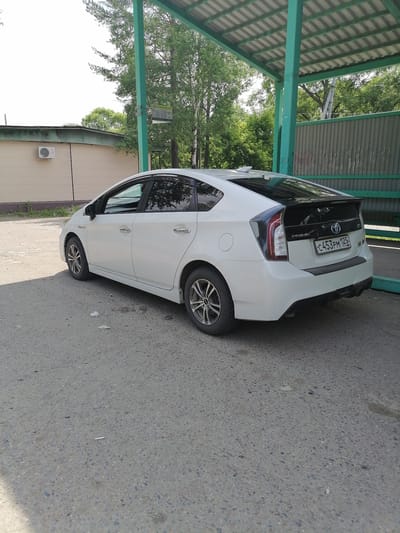 A white Toyota Prius is parked in an open area near a building with a light-colored facade and a green awning overhead. The car is viewed from the rear quarter, showing its right side and the back. The license plate reads "C453PM 125 RUS". The ground is asphalt. In the background, there are trees and a corrugated metal fence. An air conditioning unit is visible on the side of the building. The scene appears to be outdoors on a bright day. The overall impression is a street or parking area in a residential or commercial district in Novopokrovka, Russia.
