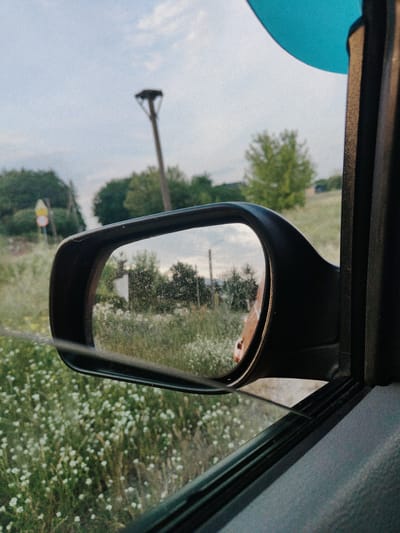 The image shows a side-view mirror of a car reflecting a rural landscape. The mirror is framed by the car's door pillar and window. The reflected scene includes a field of wildflowers, trees, and a cloudy sky. In the background, there is a tall pole with a nest on top, possibly a stork's nest. There are also some road signs visible in the distance. The overall setting appears to be a countryside road in Stanisławowo, Poland, with the time of day likely late afternoon or early evening given the soft light. The mood is serene and tranquil. The car's interior is partially visible on the right side of the frame, with a glimpse of a blue object, possibly a balloon, at the top. The mirror itself is dirty, with visible smudges and streaks.