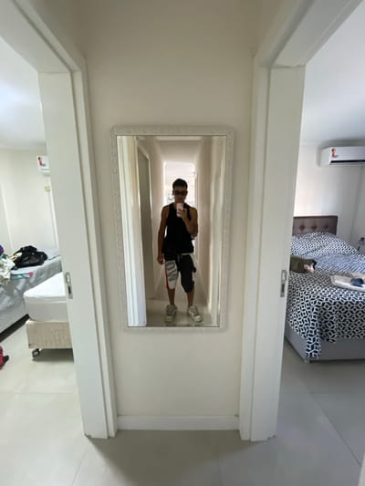 The scene is an interior hallway or common area within a dwelling in Bombinhas, Brazil. Centrally positioned on a white wall is a full-length mirror with an ornate white frame, reflecting a male individual. This individual is dressed in a black tank top, dark sunglasses, multi-colored knee-length shorts, and light-colored sneakers, and is actively taking a selfie using a light-colored smartphone held in his right hand. The floor is covered with light-toned tiles. To the left, an open doorway reveals a room containing a bed with a white mattress, upon which a black bag and clothing are situated. A white split-unit air conditioner is mounted high on the wall in this room. To the right, a second open doorway leads into another room containing a bed with a dark patterned comforter and a dark padded headboard. This room also features a white split-unit air conditioner mounted high on the wall, with light-colored textiles resting on the bed. The doorways are framed in white.