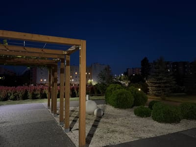 This image captures an outdoor public space at dusk in the city of Lublin, Poland. The foreground features a paved walkway made of grey brick pavers, transitioning to a bed of white gravel. A wooden pergola, constructed with thick square posts and horizontal beams, extends into the scene from the left. A vine with green leaves is growing on the pergola.

Beyond the pergola, the landscape includes neatly trimmed green bushes and two smooth, grey spherical decorative stones. Behind these, a dense row of red-flowering plants or shrubs creates a vibrant contrast against the darker background. Further in the distance, several apartment buildings with illuminated windows are visible, indicative of a residential area. Coniferous trees, including what appears to be a spruce tree, are also present. The sky is a deep, clear blue, suggesting it is nighttime. The overall scene depicts a well-maintained urban park or recreational area.