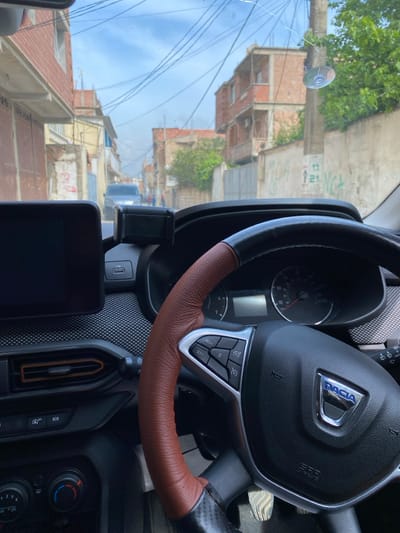 The image depicts a driver's perspective from inside a vehicle, looking out through the windshield onto a residential street in Sidi Moussa, Algeria. The foreground is dominated by the vehicle's interior. A Dacia steering wheel is prominent on the right, featuring a black central hub with a silver Dacia emblem and "SRS AIRBAG" text. The steering wheel rim is covered in a textured brown leather wrap, with a section of black carbon fiber-like material at its base. Silver-toned multi-function buttons are visible on the left spoke. To the left of the steering wheel, a black, rectangular infotainment screen is mounted on the dashboard, alongside a black universal phone holder that appears empty. Below the phone holder, a USB-A port is visible. The dashboard features a textured pattern, and an air vent with a brown/orange accent trim is located below the infotainment screen. Parts of the climate control knobs are visible at the bottom left, and the top of a silver-colored pedal is visible near the bottom right. The windshield exhibits a visible crack in the upper right quadrant, where a clear suction cup mount is also attached.

The background reveals a narrow, possibly paved, street lined with residential buildings under a partly cloudy sky. The buildings are primarily constructed of brick or concrete, with many appearing to be two or three stories high; some exhibit exposed brickwork, while others have partially rendered facades and balconies. Numerous black electrical or communication cables crisscross overhead, strung between utility poles and structures. A concrete utility pole is visible on the right side. Concrete or stucco walls line the right side of the street, displaying graffiti that includes the numbers "17," "13," "21," and letters "NGA." Green foliage is visible behind some walls and buildings. Further down the street, a dark-colored SUV or van is present, and a few indistinct human figures are visible in the distance.