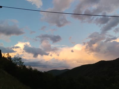 The image presents a landscape scene, likely captured at twilight, with a focus on the sky. The sky is filled with clouds of varying shapes and colors, ranging from light blue to shades of gray and tinges of orange where the sunlight appears to be reflecting. In the foreground, there is a silhouette of rolling hills covered with trees and bushes. A dark line, possibly a power line, cuts across the top of the image. Based on the prompt, the location is near Smilyan, Bulgaria. There are no visible people, actions, or interactions present in the image.