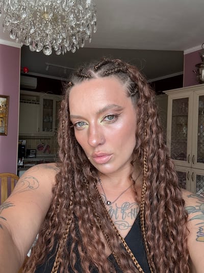 The media file is a close-up, portrait-style photograph of a person with long, wavy, brown hair styled with braids. The person is wearing makeup, including eyeshadow, and a silver necklace with a small spherical pendant. They have a nose ring. Tattoos are visible on their arms. The background suggests an indoor setting, with hints of furniture, cabinets, and a chandelier. The lighting appears to be artificial, possibly from overhead fixtures or natural light filtering in. The scene seems to be a personal space, perhaps a home, with a decorative chandelier prominent in the left foreground. The image is oriented sideways.