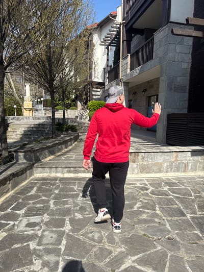 The image depicts a man standing on a stone pathway in Bozhurets, Bulgaria. He is wearing a red hoodie, black pants, and white sneakers with red accents, along with a grey baseball cap. The man has one arm raised in the air. Behind him, there is a building with a grey stone exterior and dark wooden accents. To the left, there's a bare tree and a set of stone steps leading up to a lower building. The pathway is constructed of irregularly shaped grey stones. The scene is illuminated by sunlight.