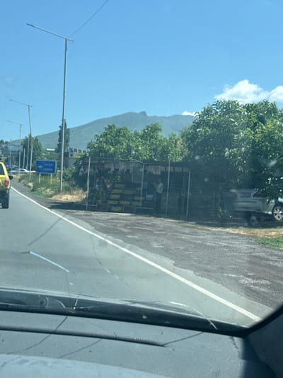 The image captures a scene on a sunny day in Karbi, Armenia, viewed from inside a vehicle through a cracked windshield. A roadside stand selling produce is visible on the right, with crates of fruits and vegetables displayed under a makeshift shelter. A person is standing inside the stand. Behind the stand, a silver car is parked, partially obscured by trees. In the background, a mountain range is visible under a clear blue sky with scattered clouds. A blue road sign with text in Armenian pointing to the left is also present near the stand. Streetlights line the road, and other vehicles are seen in the distance. The road itself is paved and has a white lane marker.