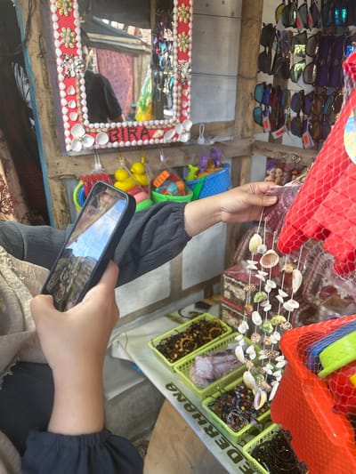 A person is holding a smartphone, capturing a photo of a string of seashell decorations. The background reveals a market stall in Bira, Indonesia, displaying various items for sale. These include sunglasses, baskets of accessories like bracelets and necklaces, and colorful plastic toys. A mirror with a seashell-adorned frame is also visible, reflecting part of the market and a person in the distance. The overall scene suggests a vibrant tourist-oriented shopping environment.