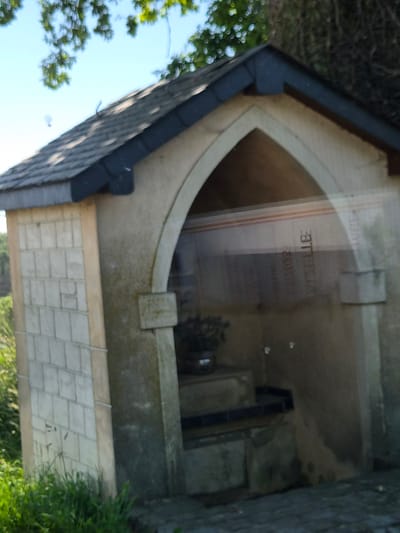 The image shows the interior of a small, stone structure with a peaked roof, likely a wayside shrine or chapel located in Lenningen, Luxembourg. Inside, there is a memorial plaque or information board visible through a glass panel. The stone structure is aged, exhibiting signs of weathering and moss growth. A stone base made up of many small blocks is built underneath the shrine. There is some green vegetation visible around the base of the structure. The view is partially obscured by light reflections on the glass.