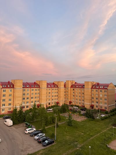 An aerial perspective captures a residential apartment complex in Kirishi, Russia, bathed in the soft, pastel hues of a sunset. The building, constructed from pale yellow brick with distinctive red-tiled roofs and visible dormer windows, stretches horizontally across the frame. Numerous white-framed windows punctuate the facade, suggesting multiple stories and apartments.
In front of the building, a paved area serves as a parking lot, containing several vehicles. These include a white van, a white SUV, a black SUV, a silver sedan, and two black sedans, all parked in an orderly fashion. Adjacent to the parking area and the building, a patch of green lawn is interspersed with several evergreen trees and a small children's playground featuring swings and colorful play structures.
Further away, more trees and green spaces are visible, contributing to the suburban feel of the scene. The sky above is a canvas of pale blue and soft pink clouds, indicative of twilight. The overall atmosphere is tranquil and domestic, depicting a typical evening in a Russian residential area.