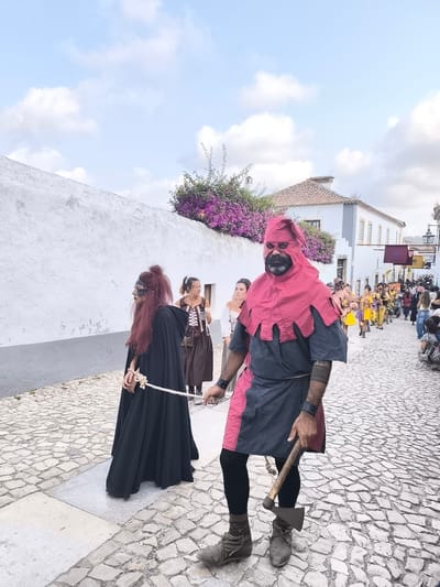 This image depicts a scene in Óbidos, Portugal, likely during a historical reenactment or festival. In the foreground, a man dressed as a medieval executioner, wearing a red hood and tunic, a dark blue undershirt, black leggings, and brown boots, holds an axe. He is walking on a cobblestone street. To his left, a person in a long black hooded robe with red and black makeup on their face, appearing to be a prisoner, is being led by a rope tied around their wrists. Behind them, other individuals in period costumes are visible. The street is lined with whitewashed buildings with red-tiled roofs, characteristic of Portuguese architecture. Purple bougainvillea flowers cascade over a white wall on the left side of the street, adding a vibrant contrast to the scene. The sky above is blue with scattered white clouds. The overall atmosphere suggests a public event with participants in historical attire.