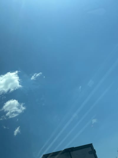 The image captures a bright, clear blue sky with scattered white clouds. The perspective suggests it was taken from inside a vehicle, looking upwards through a window, as evidenced by faint reflections and streaks that appear to be caused by the glass. In the lower right corner, the top edge of what appears to be a dark-colored vehicle or structure is visible, with a black rectangular shape that might be a cargo box or roof rack. The environment is a daytime sky, likely an outdoor setting, and the weather appears sunny and fair. There are no people or discernible actions taking place in the scene. The location is identified as Vakarel, Bulgaria.