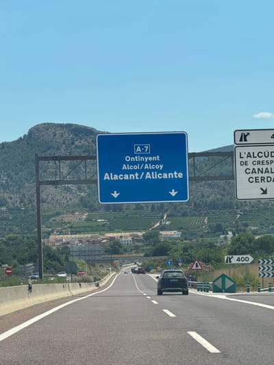 The image captures a view from a vehicle driving on a highway in Spain. In the foreground, the asphalt road is visible with white lane markings. On the right side of the road, there are safety barriers and various road signs, including one indicating "400" and another with directional arrows. Further ahead, other vehicles are traveling on the highway.

In the middle ground, a large blue overhead highway sign directs drivers to "A-7 Ontinyent Alcoi/Alcoy Alacant/Alicante" with downward-pointing arrows. To the right of this sign, another white sign is partially visible, with text that includes "L'ALCÚD" and "DE CRESP CANAL CERDA". The surrounding environment is characterized by hills covered with vegetation and scattered buildings, suggesting a semi-urban or rural setting. The sky is clear and blue. The scene appears to be set in the vicinity of Canals, Spain, based on the highway signage.