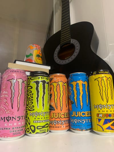 This image shows a black acoustic guitar lying horizontally, with several cans of Monster Energy drink stacked vertically on top of it. The cans are of various flavors and colors, including yellow, blue, orange, green, and pink, with visible branding and text such as "MONSTER ENERGY," "JUICED," "MANGO LOCO," "MONARCH," "VR/46," "THE DOG," "ZERO SUGAR," and "ULTRA STRAWBERRY DREAMS." One can is partially obscured by books. The background is a plain white surface, possibly a wall or countertop, suggesting an indoor setting. There are no people or animals visible in the image, and the lighting indicates it could be daytime or under artificial light. The overall composition is a still life arrangement of the guitar and the energy drink cans. The location context of Yelshanka Raz"Yezd, Russia is provided but not discernible from the visual elements of the image itself.