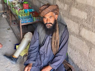 The image shows a man sitting against a wall. He is wearing a blue shirt, a brown vest, a brown turban, and a brown scarf. He has a long, white beard and is looking to the left. The man has a prosthetic leg. His left leg is bent and he is sitting against a wall. His prosthetic leg is straight out in front of him. He appears to be sitting in a store or shop. There are shelves behind him, filled with plastic bins and candy. The wall behind him is made of concrete blocks. The coordinates provided correspond to a location in the city of Panjwai, Afghanistan.