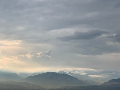 The image depicts a hazy mountainous landscape under a cloudy sky. In the foreground, a range of dark, silhouetted mountains is visible, with softer, layered mountains receding into the distance, shrouded in atmospheric haze. The sky is filled with various shades of gray clouds, with patches of lighter sky and soft sunlight breaking through in the upper left and middle portions of the image, illuminating some of the distant mountain peaks. The overall impression is one of a vast, muted natural scene. The scene appears to be located in or near Somoniyon, Tajikistan, characterized by its mountainous terrain. No people, specific objects, or actions are discernible in the image.