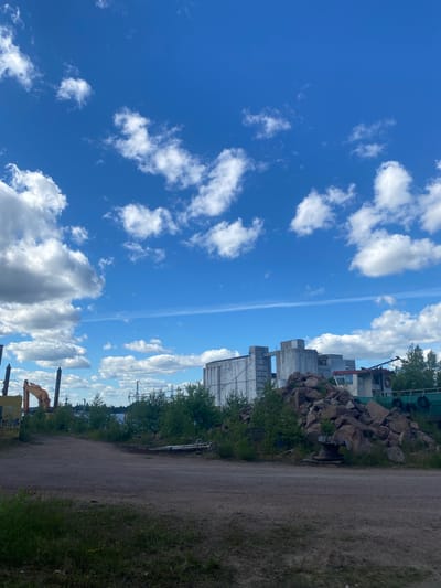 A daytime, exterior, eye-level, medium shot captures a gritty industrial landscape in Kotka, Finland, under a bright blue sky filled with fluffy white clouds. In the foreground, a dirt path cuts across the scene, with patches of green grass and small white wildflowers visible along its edges.
To the left, a yellow excavator, a common piece of construction equipment, stands partially visible, its arm extended. Further back, a line of dark, vertical poles rise from the ground, some appearing to be dock pilings or industrial structures. Behind these, lush green trees and vegetation form a dense line along the horizon.
Dominating the mid-ground and extending into the background are industrial buildings. A large, multi-story concrete structure with a grey, weathered facade stands prominently on the right. To its left, another connected concrete industrial building is visible. A barge or ship, painted green with black buoys along its side, is partially obscured by a large pile of rough, brown rocks situated in front of the industrial buildings. The pile of rocks is quite substantial, suggesting a recent or ongoing material handling operation. A wooden cable spool rests on the ground near the base of the rock pile.
The overall impression is one of an active, if somewhat unrefined, industrial port or yard, with evidence of construction and maritime activity. The bright sky and scattered clouds suggest fair weather conditions.
