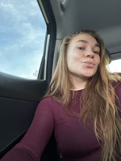 A woman is sitting in the passenger seat of a vehicle, looking at the camera. She has long, blonde hair and is wearing a maroon long-sleeved shirt. The background shows the sky through the car window and the interior of the car. The scene is in Ryabinovka, Russia.