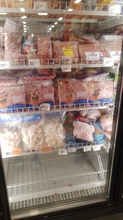 The image displays the interior of a commercial upright display freezer within a retail establishment located in Kahawa Sukari ward, Kenya. The freezer features a glass door and multiple white wire shelves.
The top visible shelf contains various vacuum-sealed plastic packages of what appears to be minced meat, some labeled "Mince" or "FROZEN," with price tags reading 379/-, 925/-, and 579/-. The second shelf from the top is filled with numerous clear plastic packages of "Farm Fresh" branded minced beef, with visible labels stating "Minced Beef Mince" and "Frozen Beef Mince," accompanied by price tags such as 300/- and 339/-.
The third shelf from the top holds multiple clear plastic bags of "Shrimps," visible on the left, alongside other packages containing white, peeled pieces resembling garlic or similar vegetables, priced at 530/- and 100/-. On the right side of this shelf are several opaque plastic-wrapped items, consistent with whole or processed poultry, including one package branded "CAPIA," with price tags of 680/-, 290/-, and 715/-.
The bottom two shelves are empty, revealing only the white wire rack structure. Reddish-brown residue or staining is present on the white plastic base of the freezer, below the lowest shelf. No individuals or active interactions are visible.