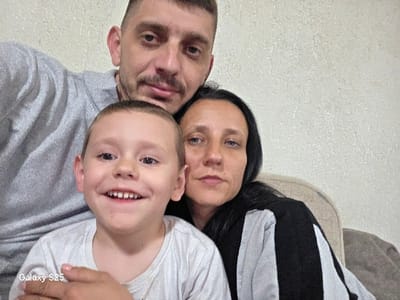 This image displays three individuals in an indoor setting in Karnalovo, Bulgaria. In the foreground, a young male child, approximately 5-7 years old, with short light brown hair and a white t-shirt, smiles broadly, revealing his teeth, and looks directly at the camera. A hand is visible resting on his right upper arm. Behind and slightly to the right of the child stands a male adult, estimated to be in his 30s-40s, with short dark hair, a trimmed beard, and a grey hooded sweatshirt. He maintains a neutral expression while looking at the camera. To the right of the child and adjacent to the male adult is a female adult, also appearing to be in her 30s-40s, with long dark hair and a black and white striped garment. She too looks directly at the camera with a neutral expression. The background features a light-colored, textured wall. A portion of a beige upholstered chair is visible to the right of the female adult. The bottom left corner of the image contains the text "Galaxy S25."