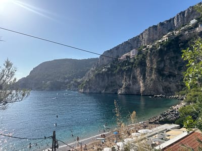 This image presents a panoramic daytime view of Mala Beach in Cap-d'Ail, France. The scene is illuminated by bright sunlight, indicated by a strong lens flare in the upper left, against a clear blue sky. The central feature is a deep blue-green bay, with lighter turquoise water visible closer to the shore. Numerous individuals are dispersed within the bay, swimming or wading. Several small boats are present, anchored or moving across the water.

The bay is framed by prominent, steep, rocky cliffs, densely covered with green vegetation. On the right side, multiple multi-story residential buildings and villas, characterized by light-colored facades and red-tiled roofs, are constructed into and along the cliff faces. A densely forested hill forms the left side of the bay.

At the base of the cliffs, a narrow, crescent-shaped pebble beach is active with numerous people. An organized section on the right portion of the beach features rows of dark sun loungers and umbrellas. Closer to the foreground, white beach umbrellas and tables indicate beachside establishments. The immediate foreground includes blurred green foliage on both sides and a prominent dark horizontal wire traversing the upper-middle section of the image, with a vertical support structure partially visible below it. A portion of a red-tiled roof is present in the lower right corner.