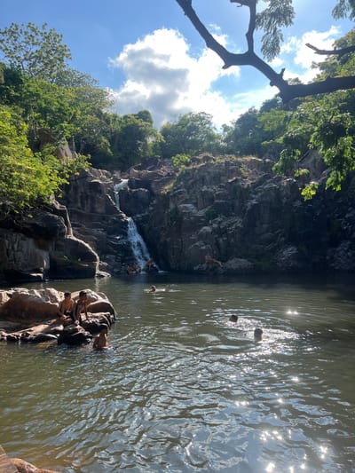 The image depicts a natural landscape with a waterfall cascading down a rocky cliff into a pool of water. Several individuals are present in and around the water. In the foreground, three young males are seated on large rocks on the left side of the frame. To the right of them, another young male stands in the water. Further back, near the base of the waterfall, a group of people are seated on rocks, with some partially submerged in the water. Two individuals are swimming in the pool in the middle ground. The environment is lush with green trees and vegetation covering the rocky terrain and surrounding hills. The sky is blue with scattered white clouds, and sunlight reflects off the surface of the water. The location is identified as a waterfall in Vĩnh Hải, Vietnam.