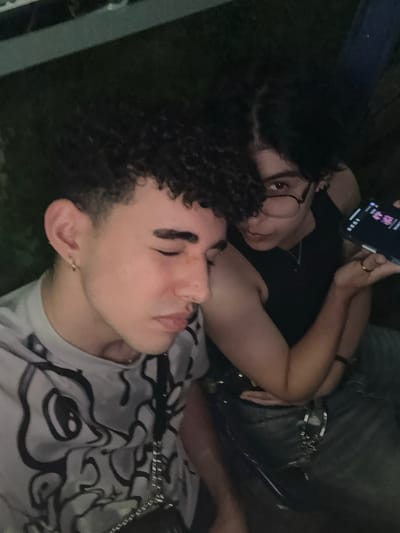 Two people are close together. One person is on their back, eyes closed. They have dark, curly hair and a light-colored patterned shirt. The other person is propped up, looking at the camera while holding a phone. They have dark hair and glasses and are wearing a black shirt. A chain belt is visible. The photo is taken from an overhead angle.