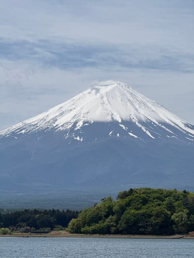 A majestic landscape photograph features the snow-capped Mount Fuji on a bright, partly cloudy day. The iconic volcanic peak, covered in white snow and ice, dominates the left side of the frame, its rugged slopes contrasting with the clear blue sky. Wispy, delicate clouds drift across the upper part of the sky, adding texture above the mountain.
Below and to the right of Mount Fuji, a lush, dense green forest forms a prominent horizontal band across the mid-ground. This verdant tree line borders a large body of water, likely a lake, which occupies the lower right section of the image. The lake's surface appears calm with subtle ripples, reflecting the sky. In the far distance, behind the forest, faint, hazy blue mountain ranges recede into the horizon, adding depth to the panoramic view.
The scene depicts a serene natural environment during daytime, with clear visibility. The location cues, especially the distinct appearance of the mountain, strongly suggest the area around Fujikawaguchiko, Japan. There are no people, specific activities, or visible text in the image. The overall impression is one of peaceful grandeur and natural beauty.