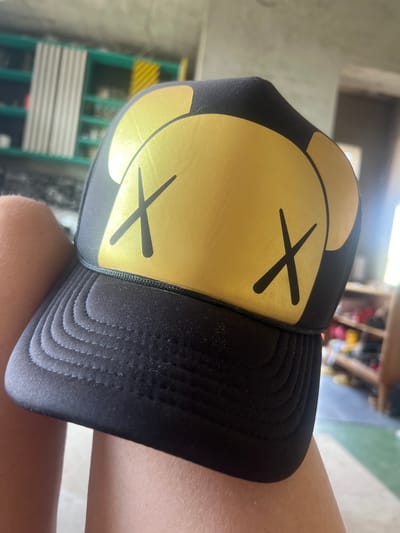 A close-up, angled shot captures a black trucker hat with a large, gold, cartoonish face design. The face features two "x" marks for eyes and a round outline for the head. The hat appears to be resting on someone's leg, with the brim visible and detailed stitching. The background is blurred, showing an indoor setting with shelves and what looks like a ladder. The lighting suggests daytime, with natural light coming from an unseen source. The overall mood is casual and relaxed. No visible text is present on the hat or in the scene.